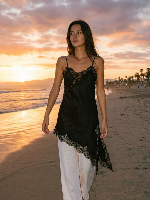 Woman in black lace top and white pants walking on a beach at sunset.