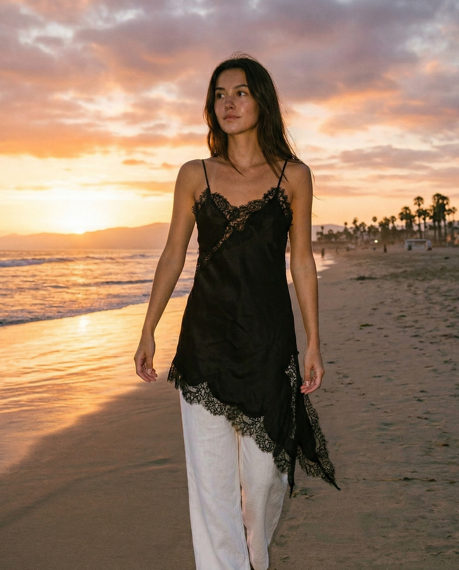 Woman in black lace top and white pants walking on a beach at sunset.